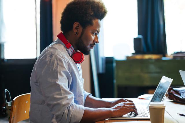 A person working on a laptop, wearing red headphones around their neck, with a takeaway cup on the desk next to the laptop.