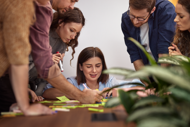 A group of colleagues appears to be in a workshop, all leaning over a table covered with Post-it notes.