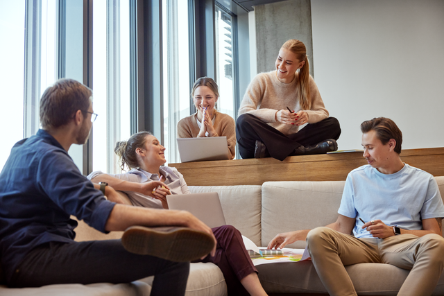 Five colleagues are sitting and talking in a lounge area of an office, with comfortable seating and a casual atmosphere.