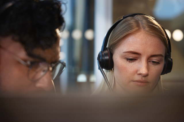 Two people wearing headsets and appear to be working at computers.