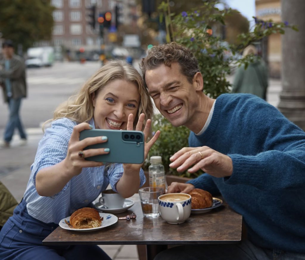 Two people sit at an outdoor table with coffee and pastries. They are smiling and waving at a phone one of them is holding, suggesting they are on a video call.